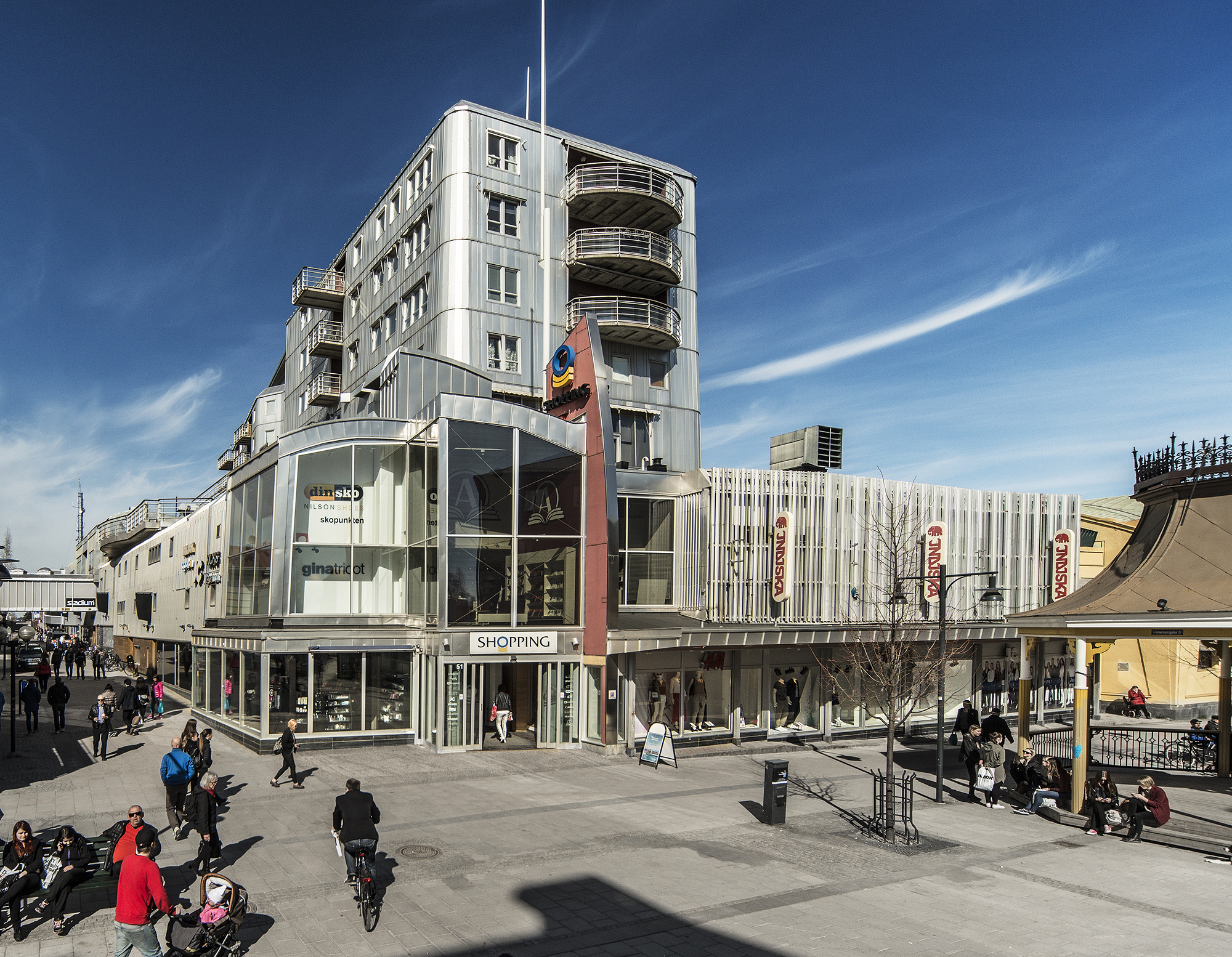 'Shopping galleria i Luleå. Det är sommar och blå himmel med slöjmoln. Människor går och cyklar på gågatan utanför.'
